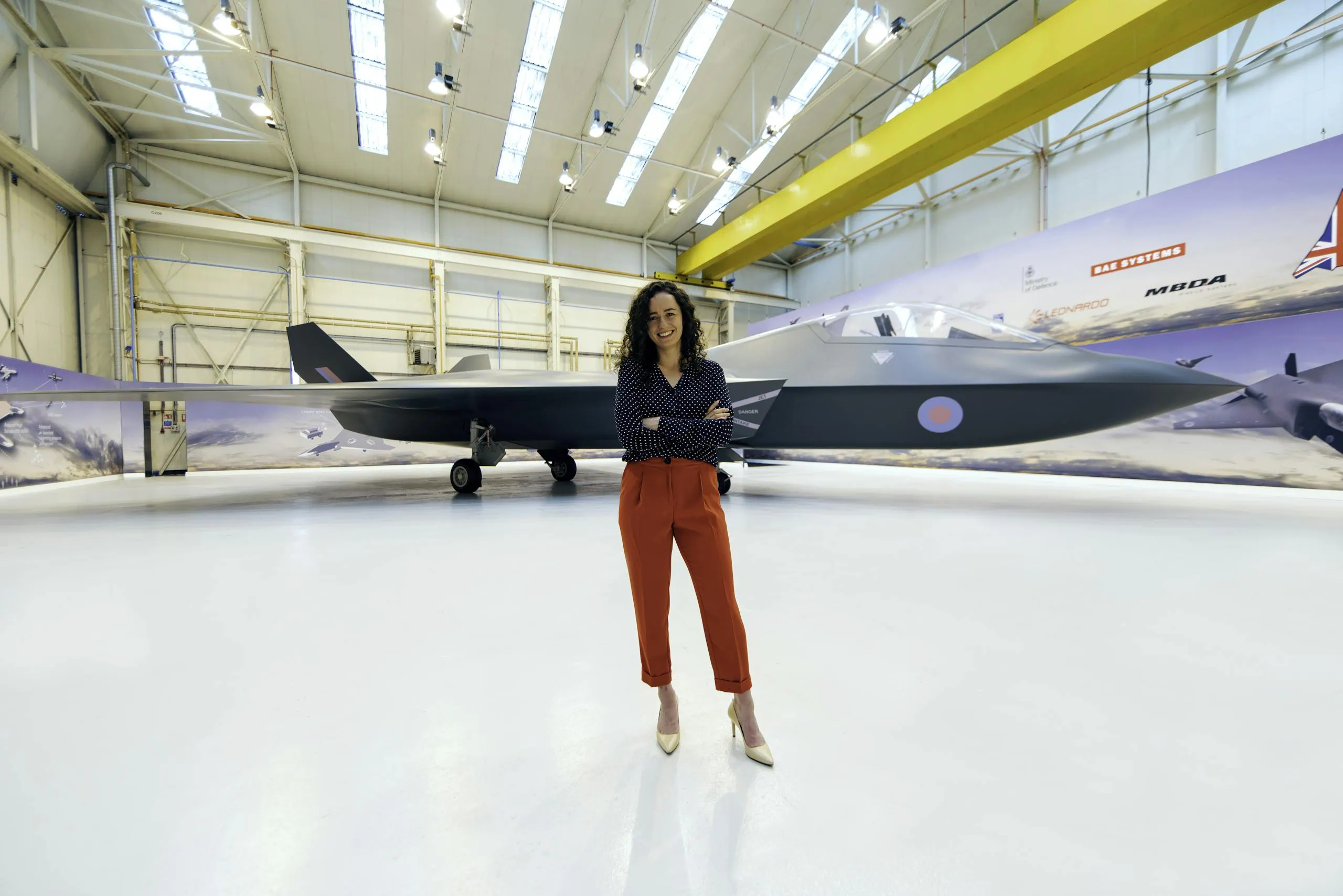 A female engineer stands confidently in front of a modern fighter jet in a hangar.